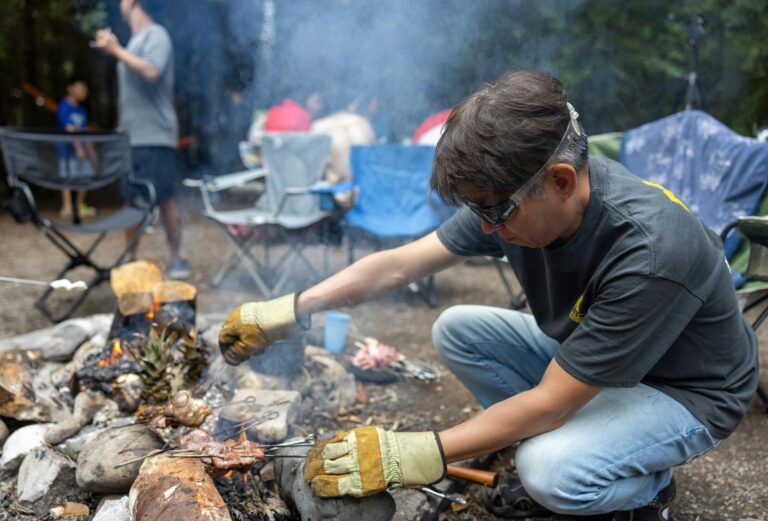 A man grilling meat over a smoky campfire in an outdoor picnic setting with others relaxing nearby.